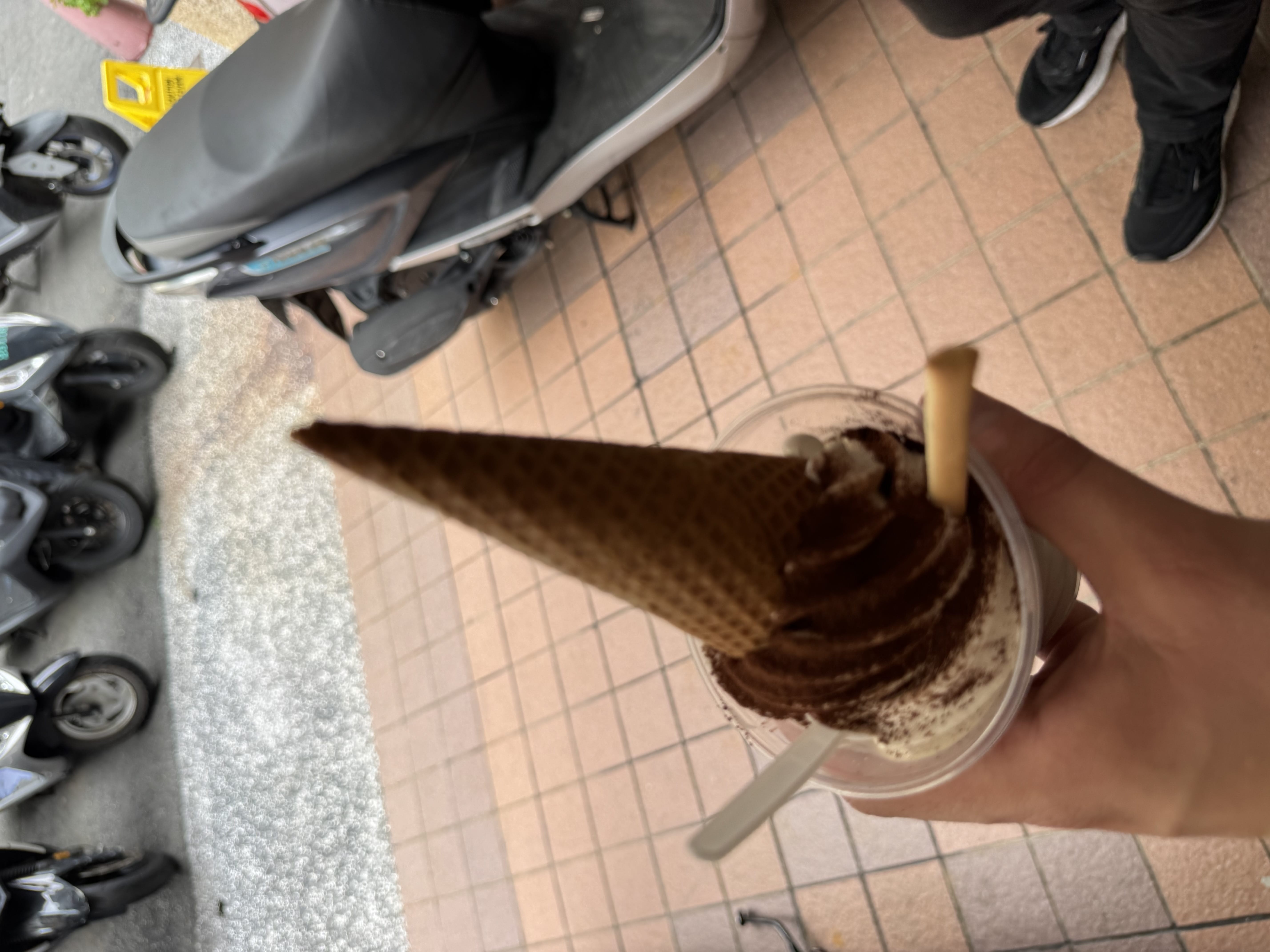 Soft-serve ice cream topped with cocoa and an upside-down cone in a plastic cup; scooters parked in the background.