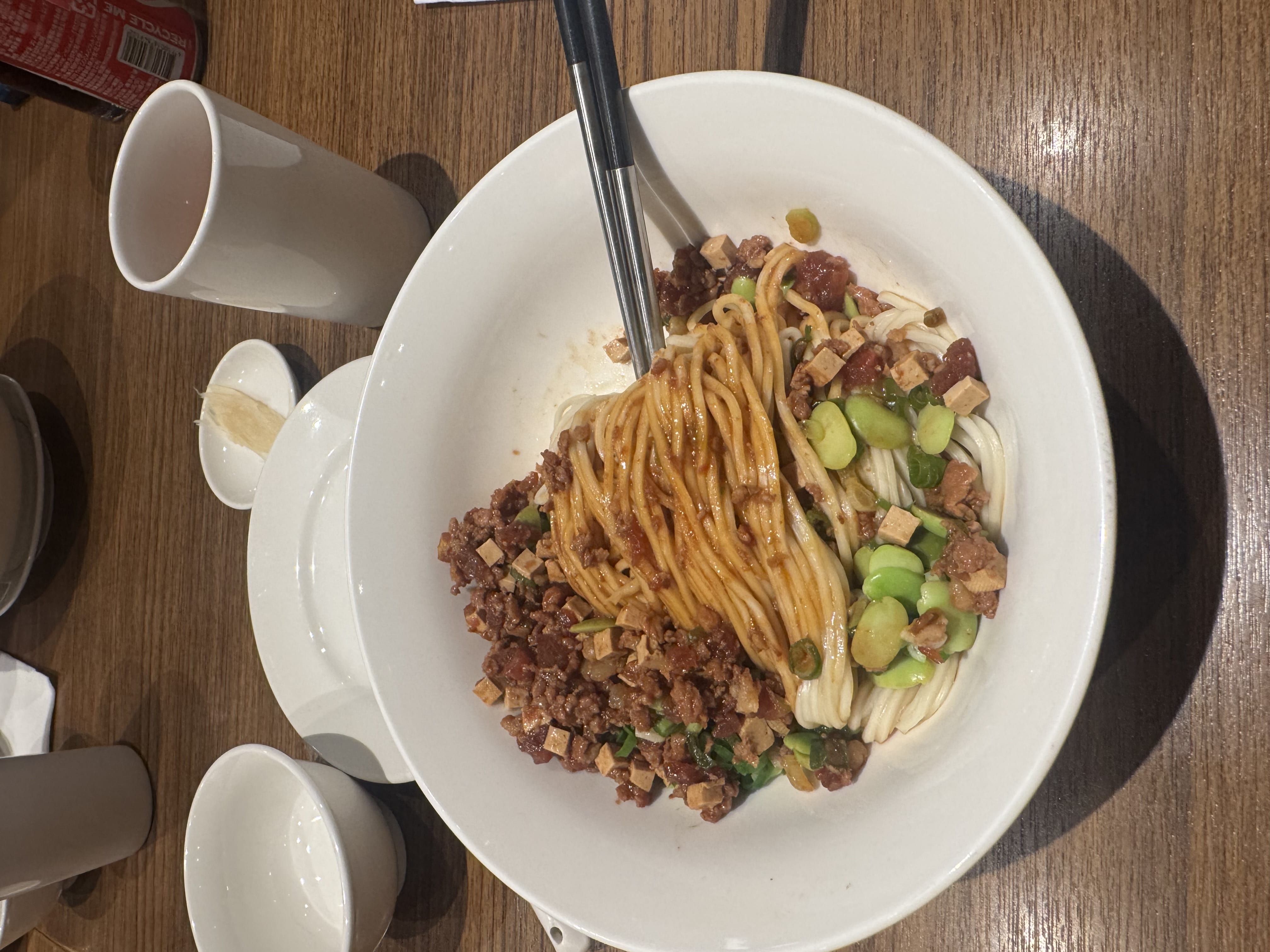 Bowl of noodles topped with minced pork, and green vegetables on a wooden table.