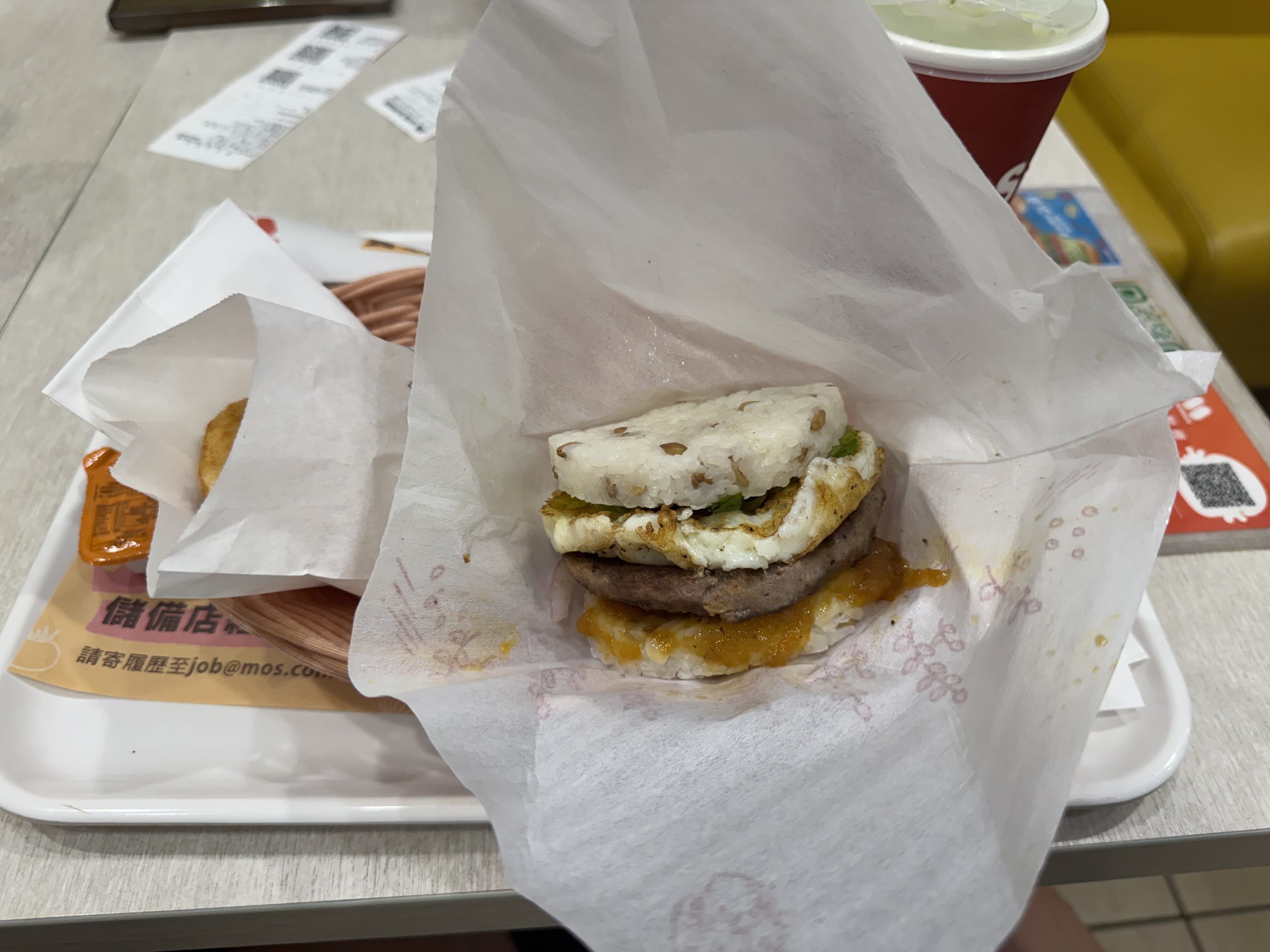 MOS Burger rice-bun sandwich with egg and patty, wrapped in paper on a tray.