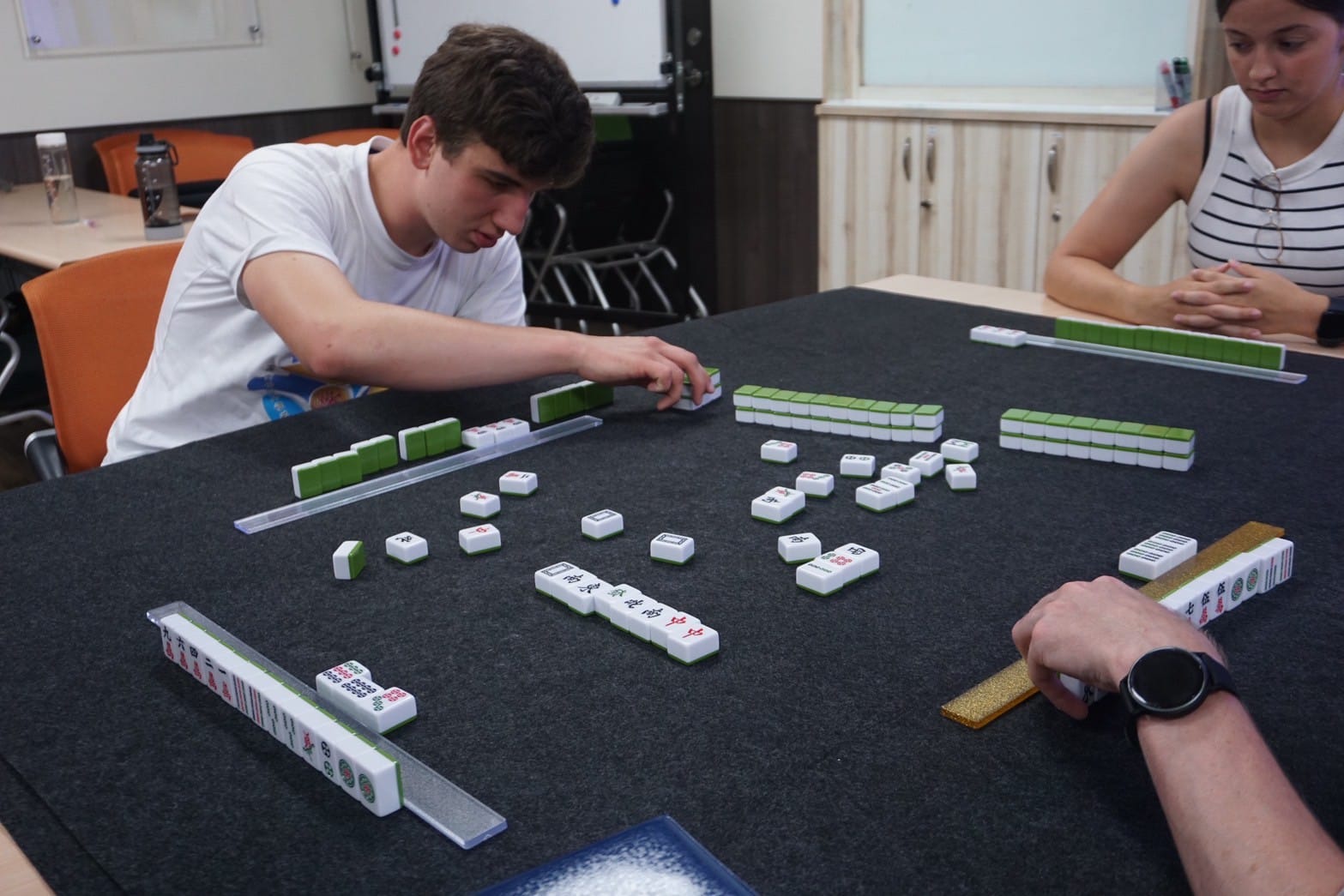 Students around a table playing mahjong; tiles arranged on a felt surface.