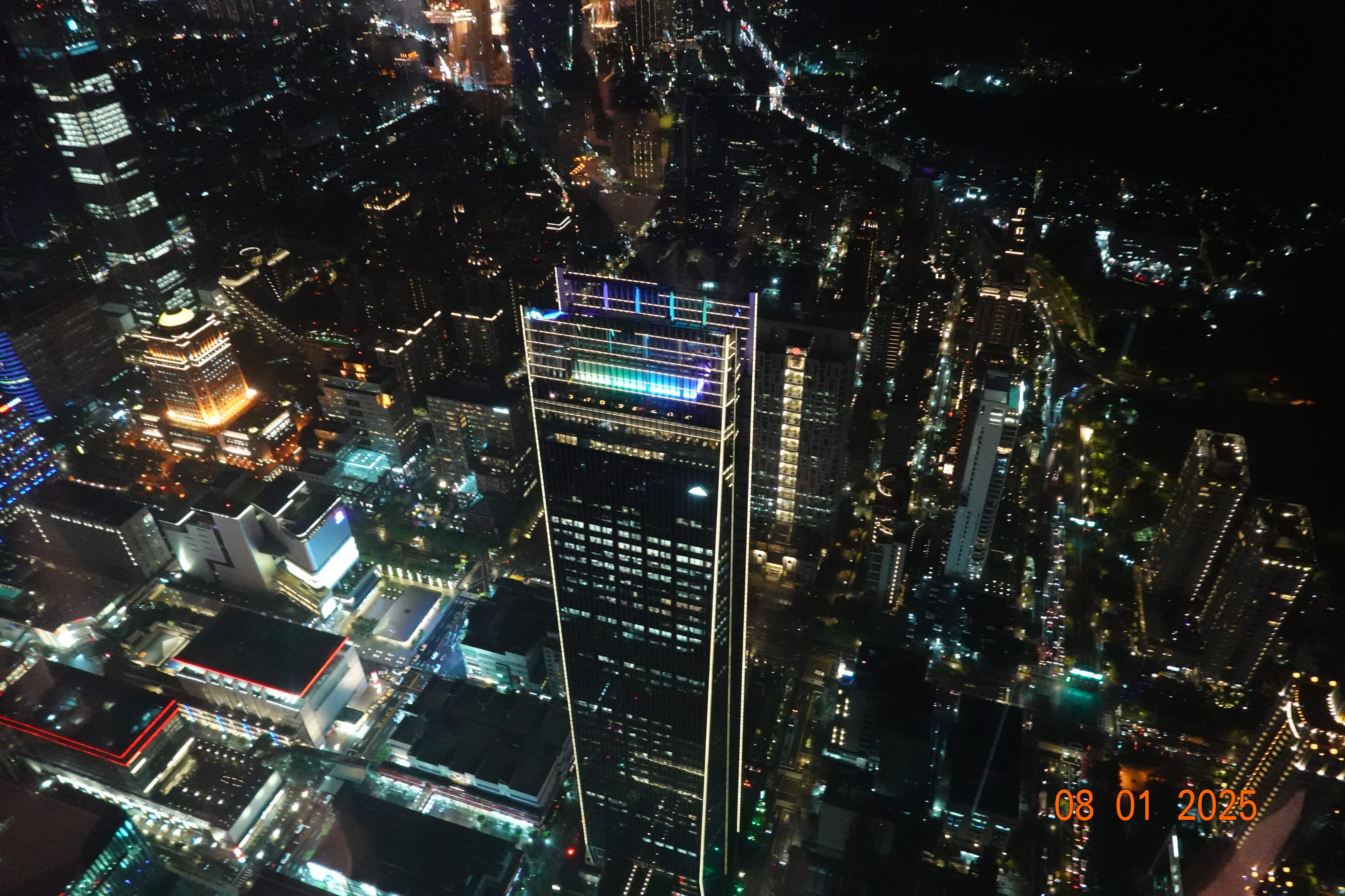 Night aerial of Taipei featuring a tall tower with a colorful illuminated crown amid dense city lights; date stamp 08-01-2025. loading=