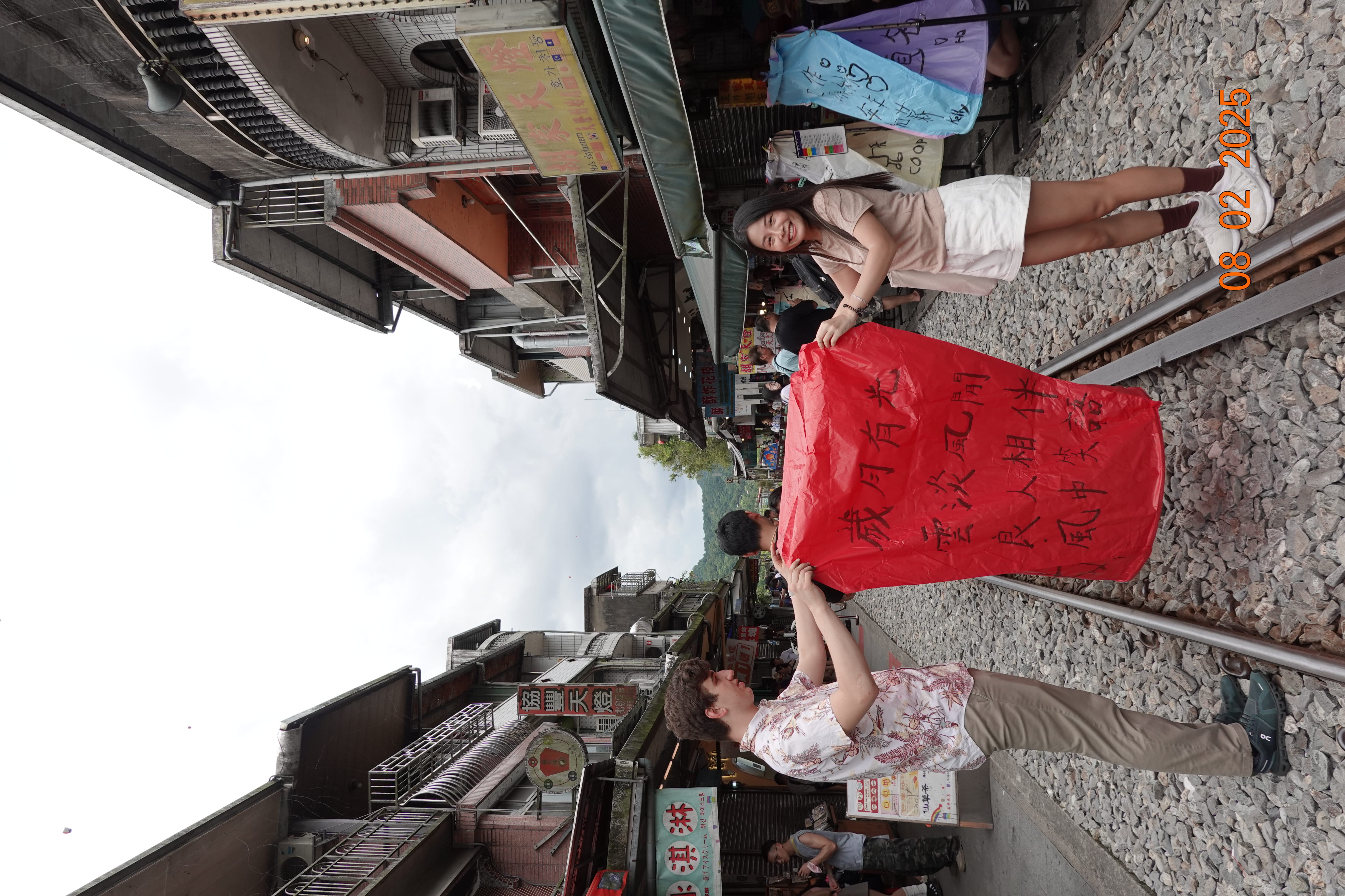 Two people holding a red sky lantern with handwritten Chinese characters on train tracks in a market street; 08-02-2025.
