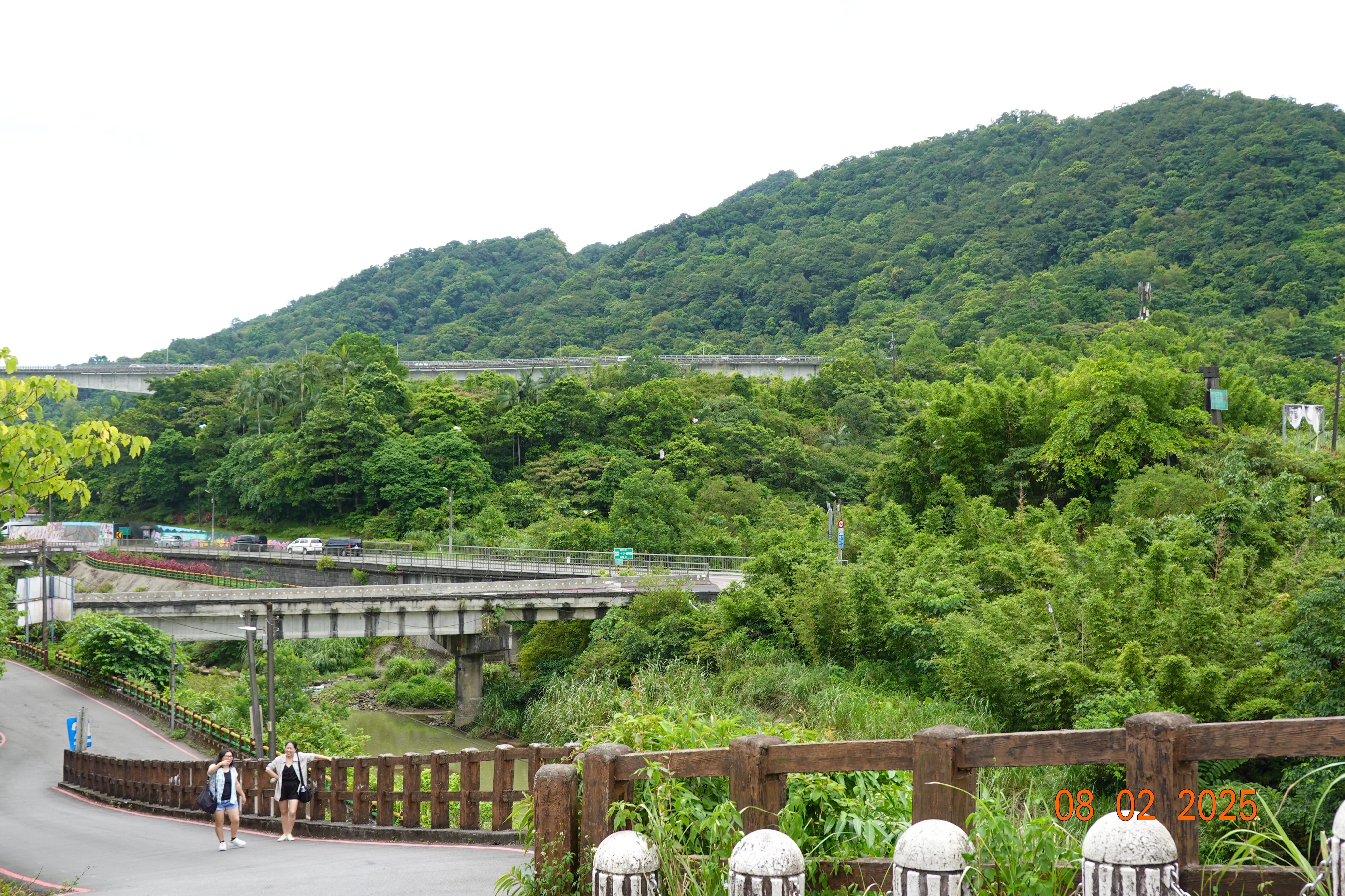 Green hillside landscape with layered roads and bridges crossing a small river valley; 08-02-2025.