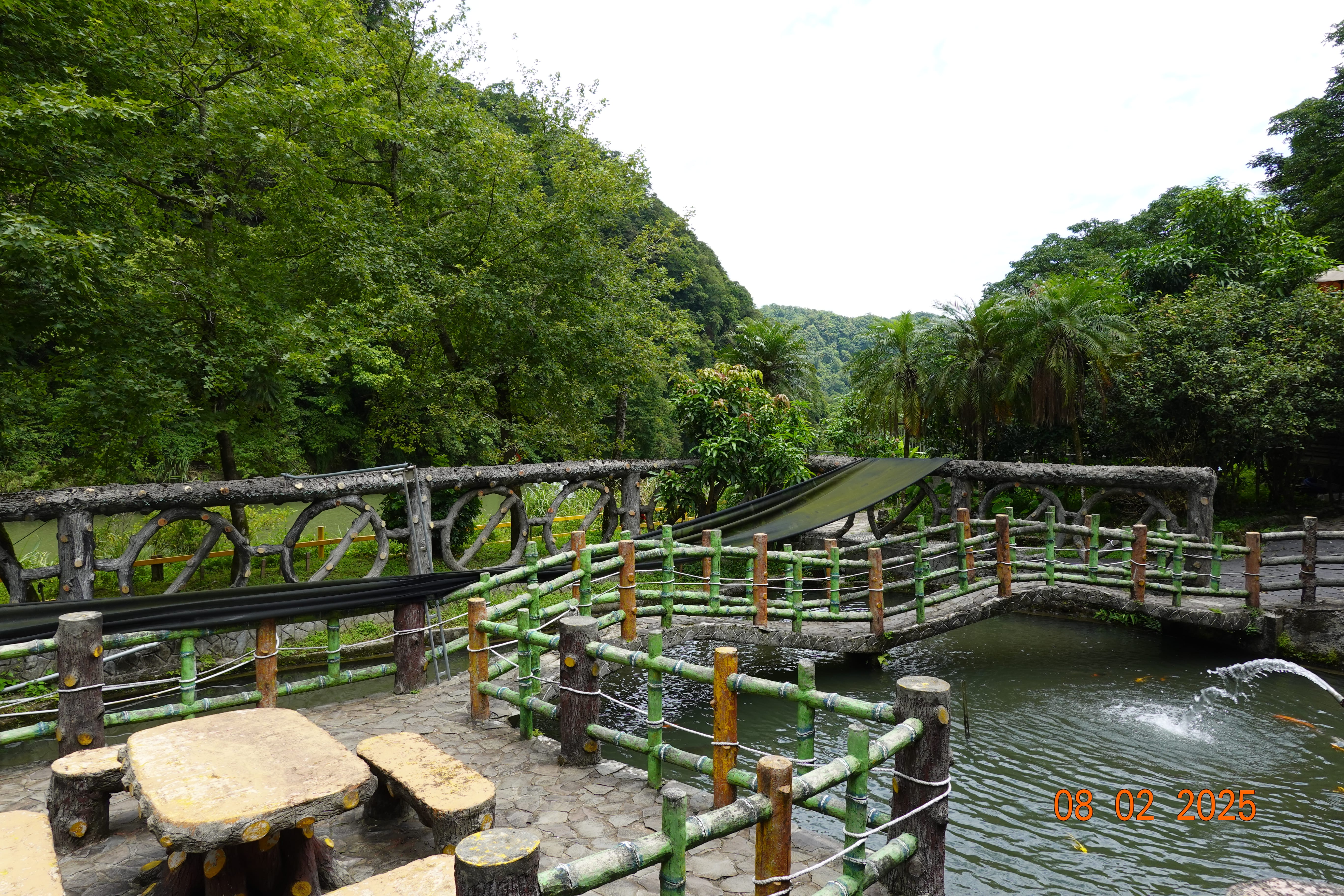 Garden pond with rustic bridges and bamboo-style railings, stone tables, and palms against forested hills; 08-02-2025.