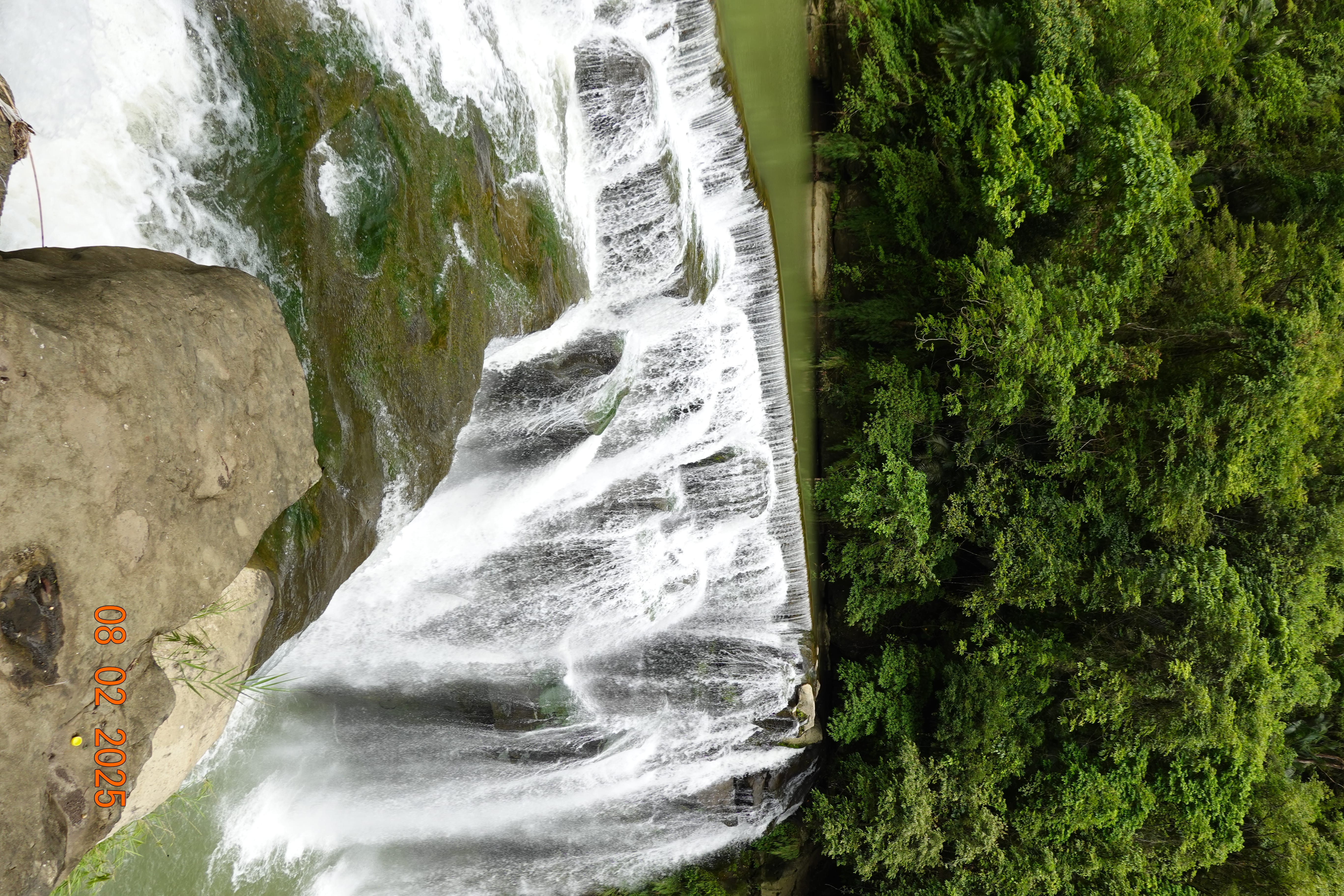 Wide tiered waterfall spilling over green-tinted rock into a pool, with lush forest backdrop; 08-02-2025.
