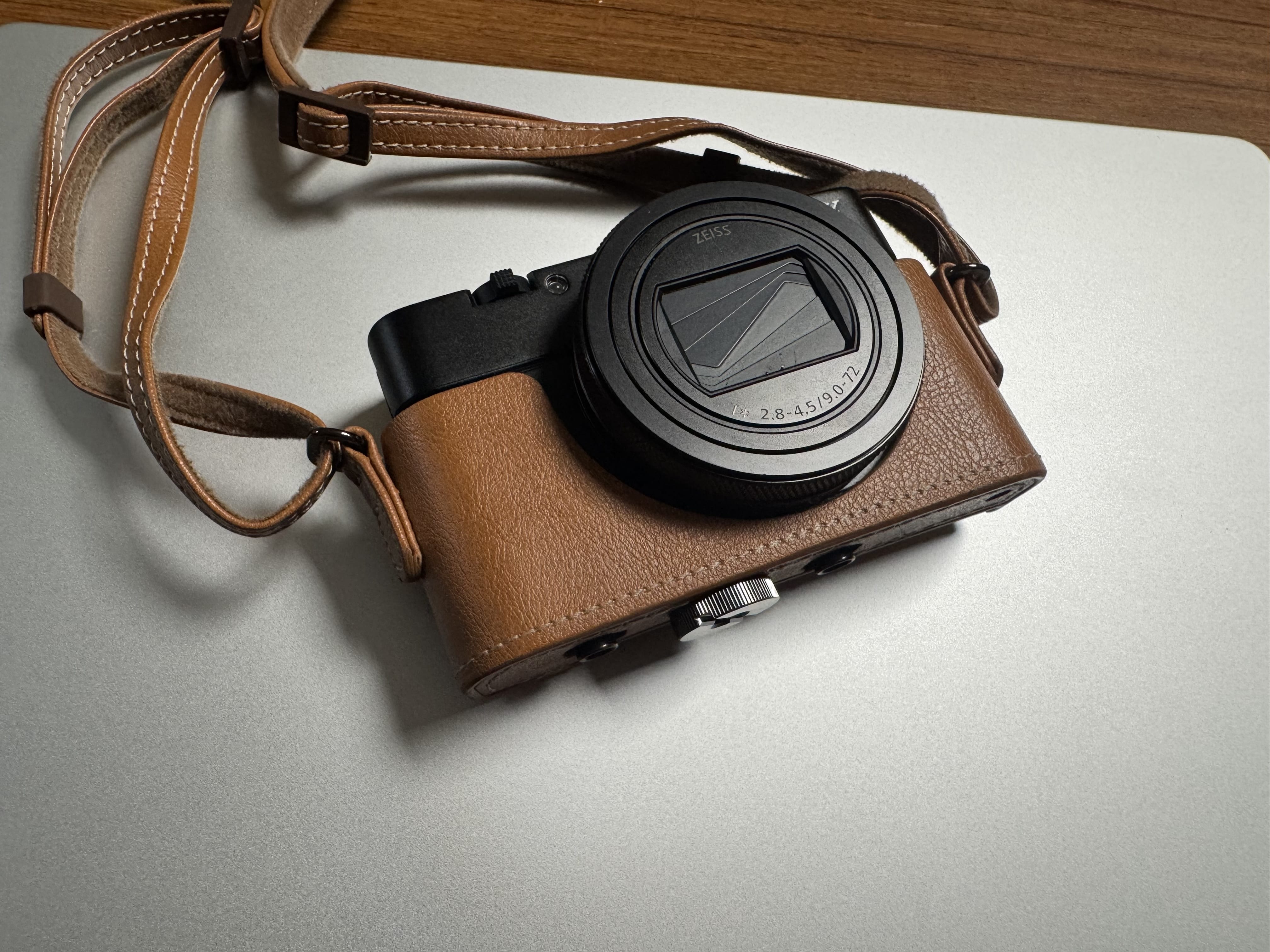 Sony RX100 VII in a tan leather half-case resting on a silver laptop, lens closed and strap looped.