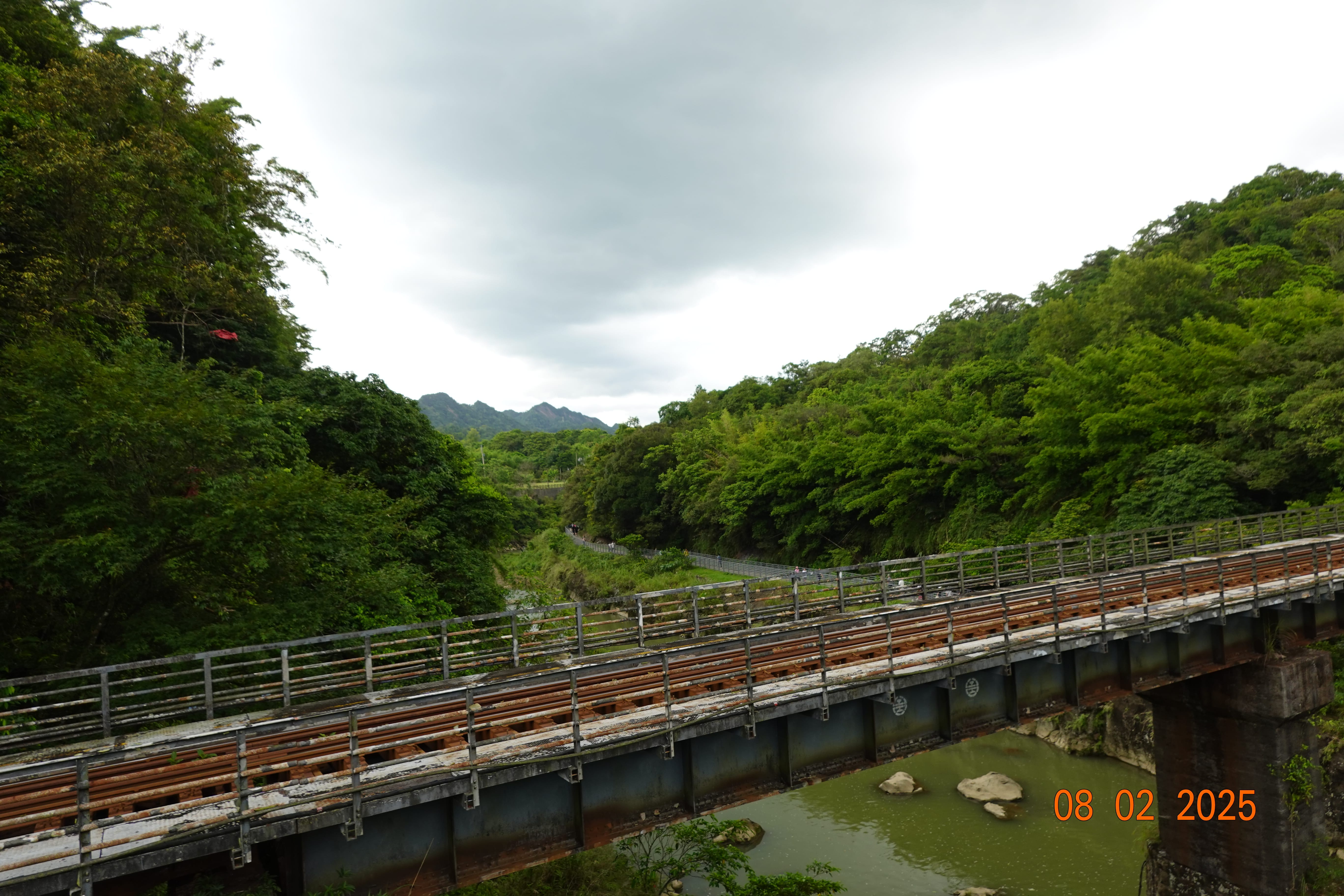 Old steel railway bridge crossing a green river gorge amid lush hills under an overcast sky; 08-02-2025.