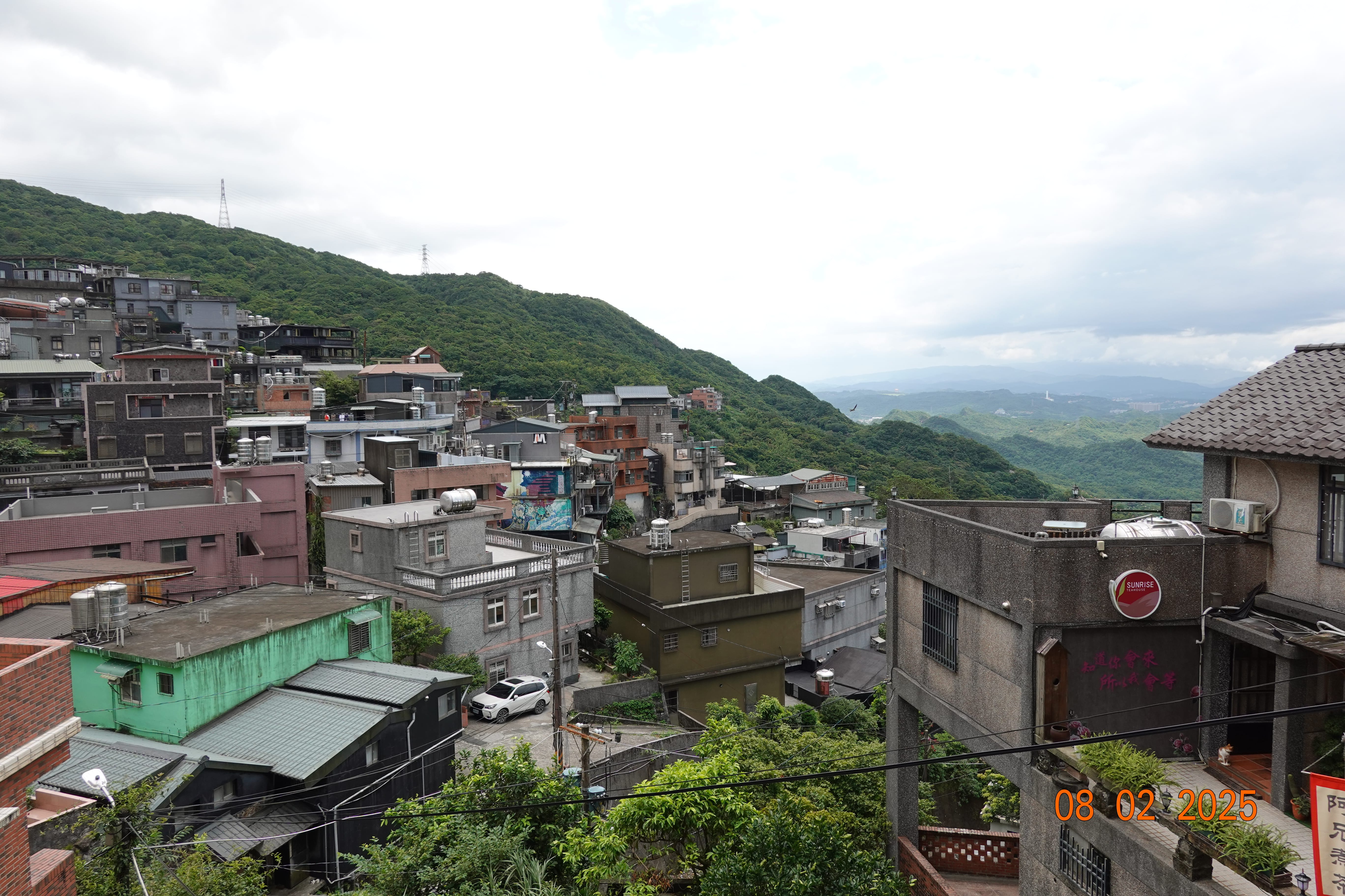Hillside town of Jiufen—stacked concrete homes and rooftop tanks stepping down toward misty green mountains; 08-02-2025.