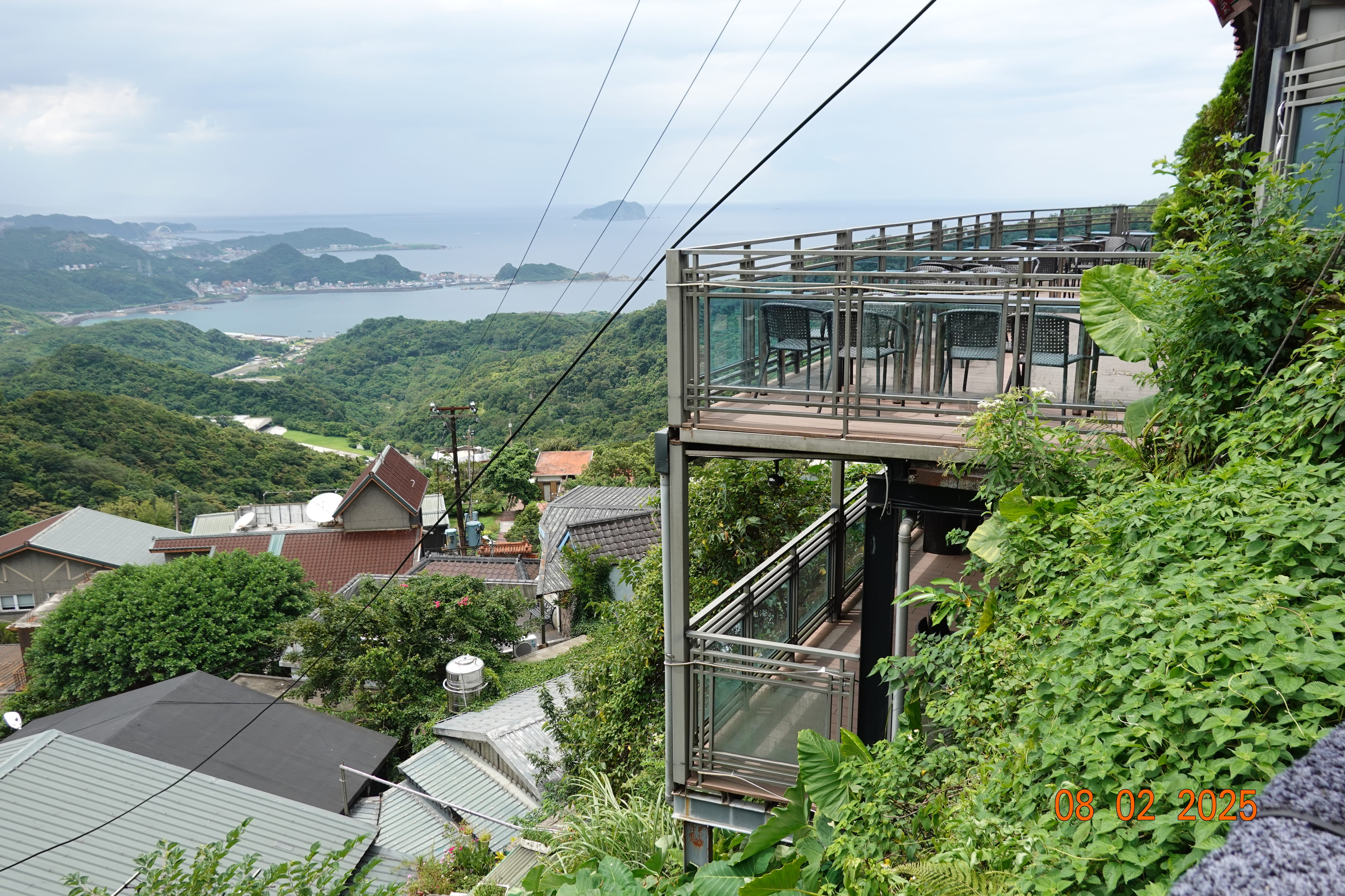 Cliffside terrace café with empty tables overlooking green hills and Taiwan’s northeast coast with offshore islets; 08-02-2025.