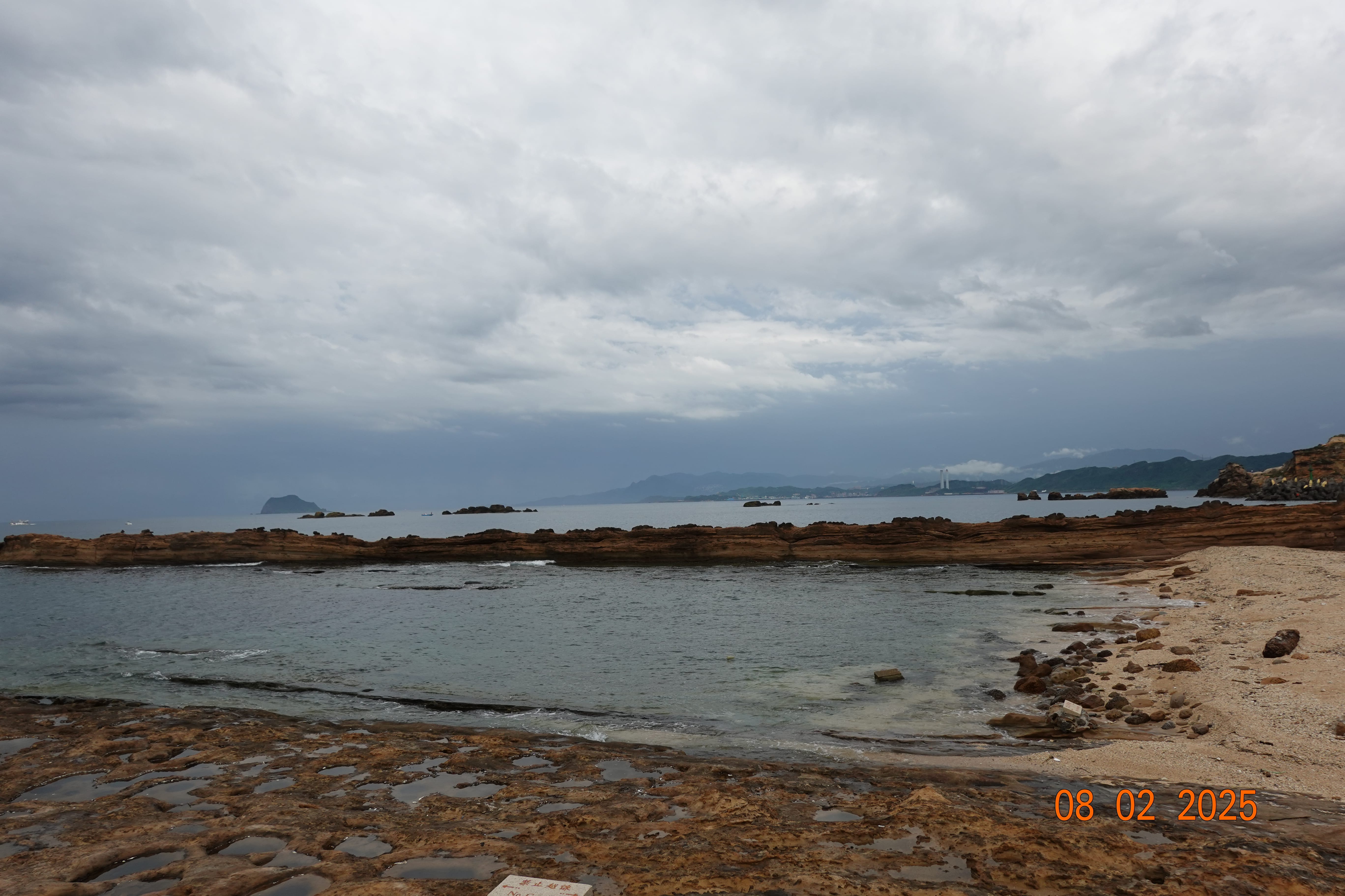 Rocky shoreline with shallow tidal pools and a long brown reef facing distant headlands and an offshore islet; 08-02-2025.