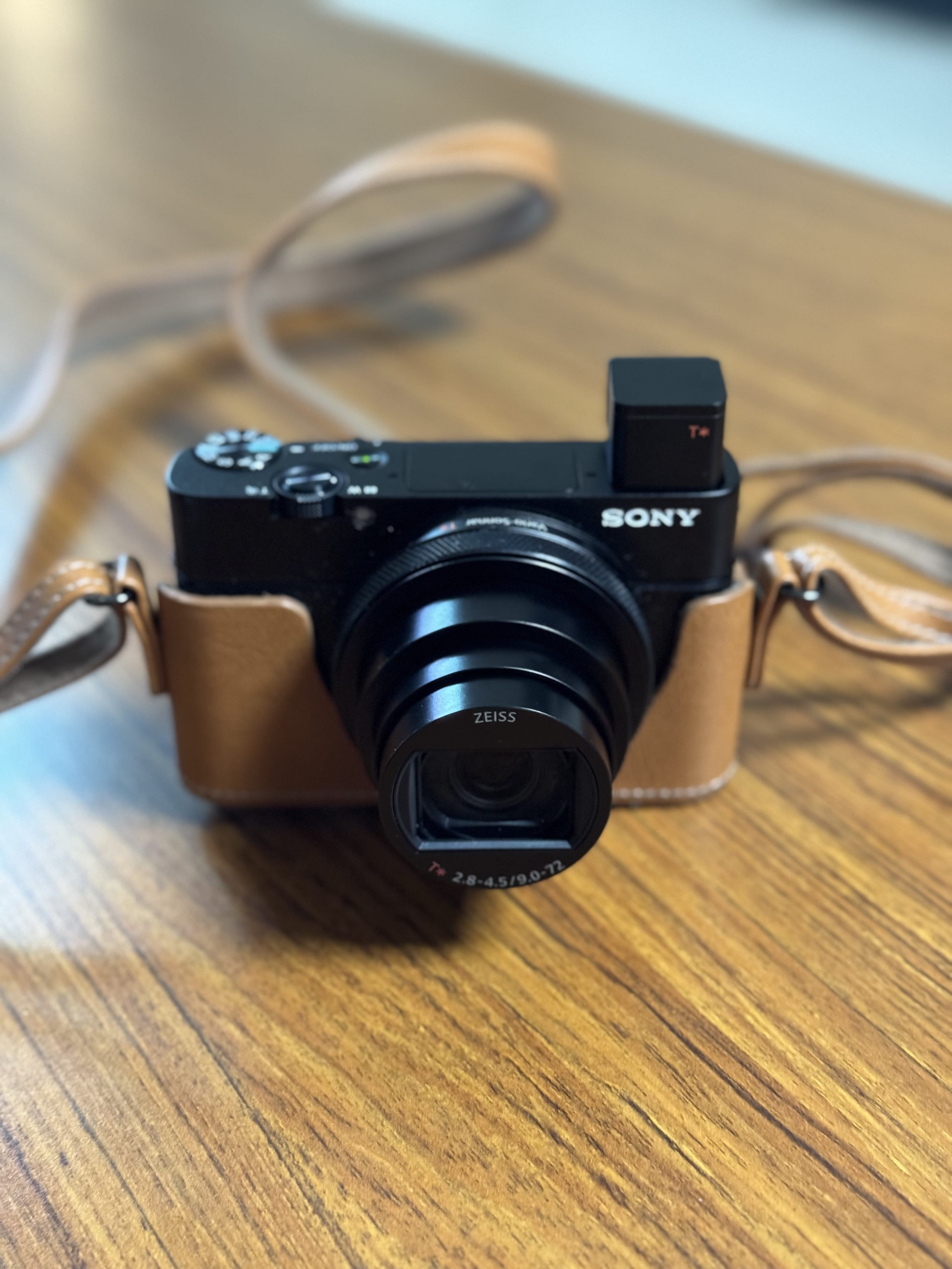 Sony RX100 VII compact camera with the pop-up electronic viewfinder raised and the lens extended, in a tan leather half-case on a wooden table.