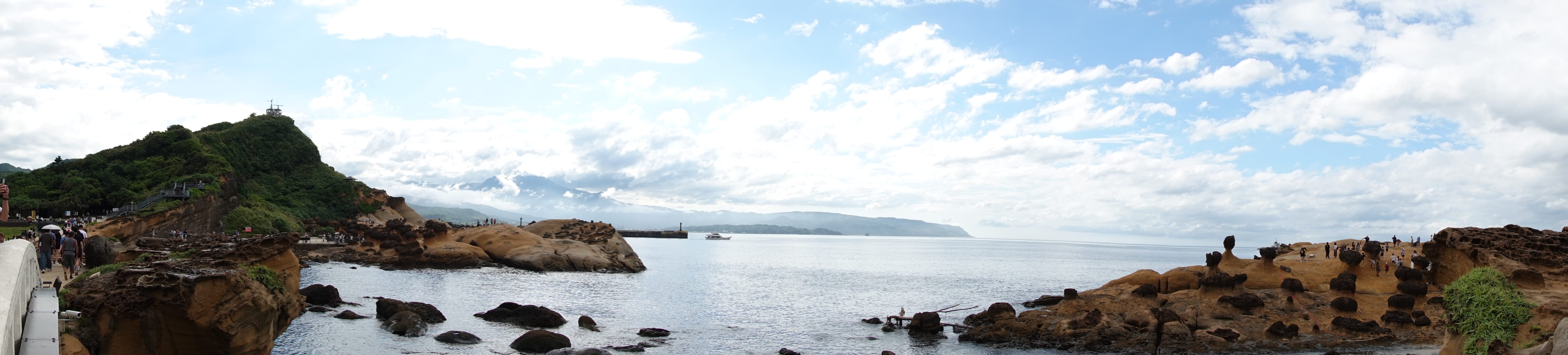 Panoramic view of Yehliu’s sculpted sandstone coast and calm sea under a sky of scattered clouds, visitors exploring along the rocks.
