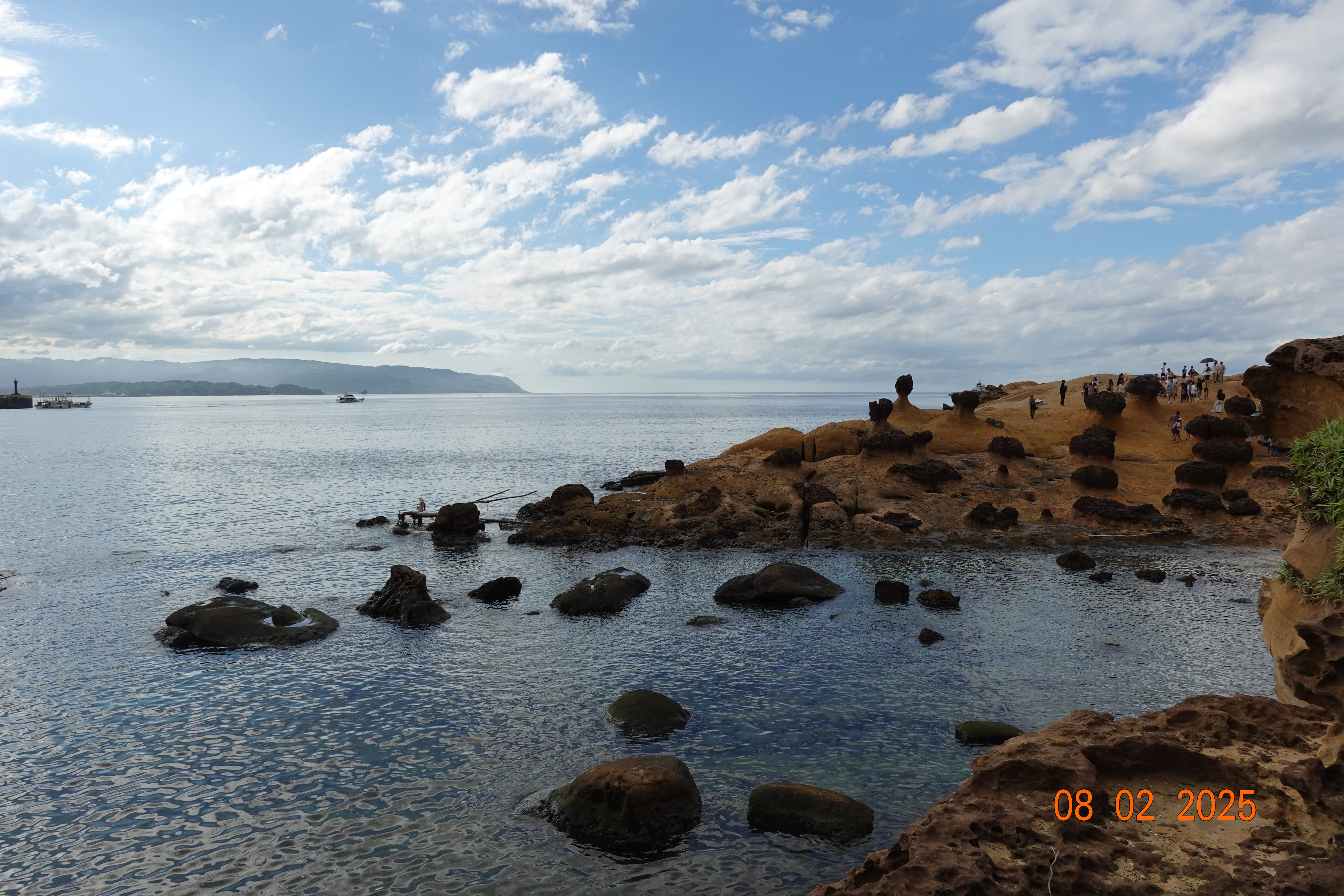 Rocky tidal pool bay and honeycomb sandstone hoodoos at Yehliu, people on the ridge and boats offshore; 08-02-2025.