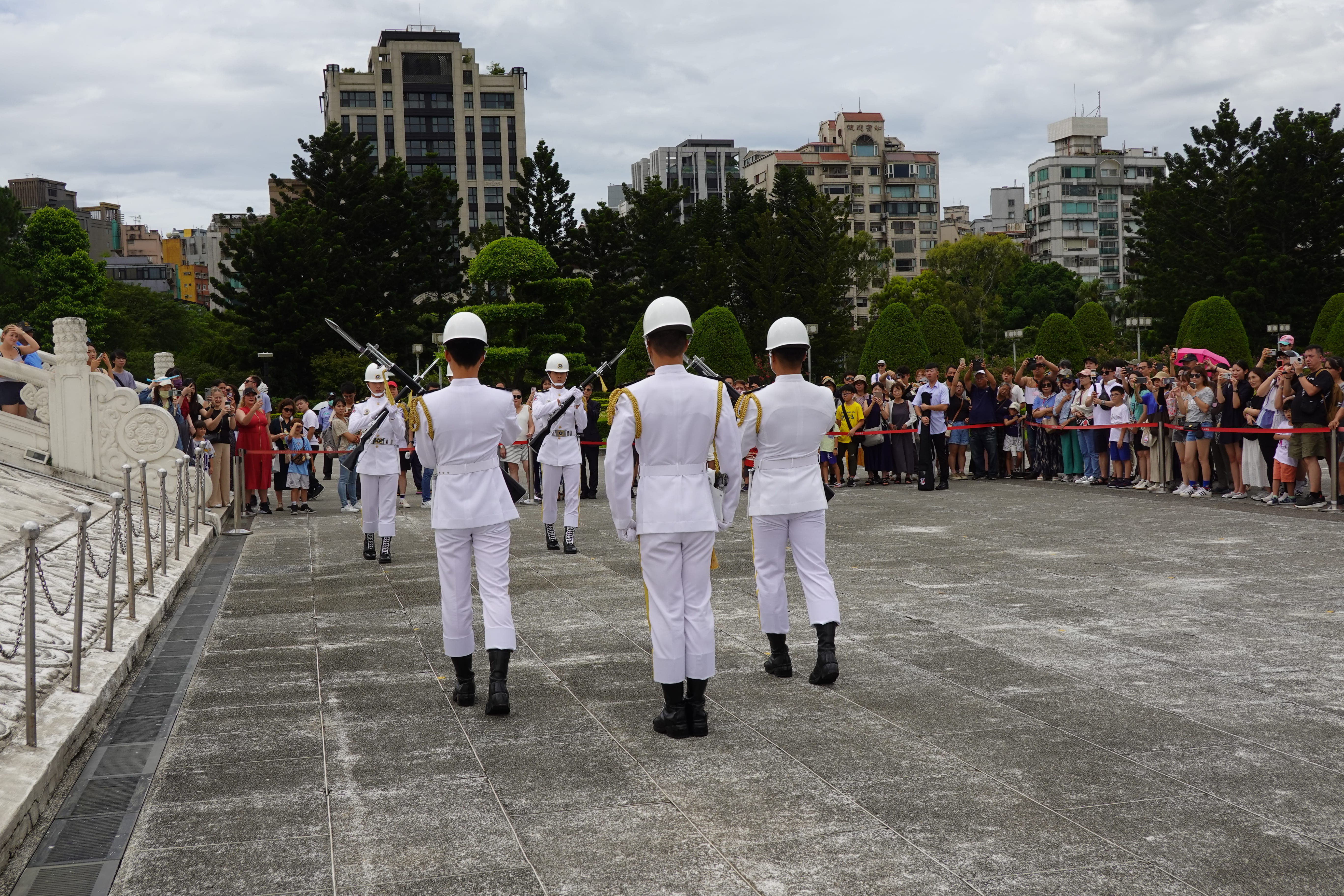 White-uniformed honor guard performing a rifle drill during a ceremonial change in a memorial plaza as crowds watch behind ropes.