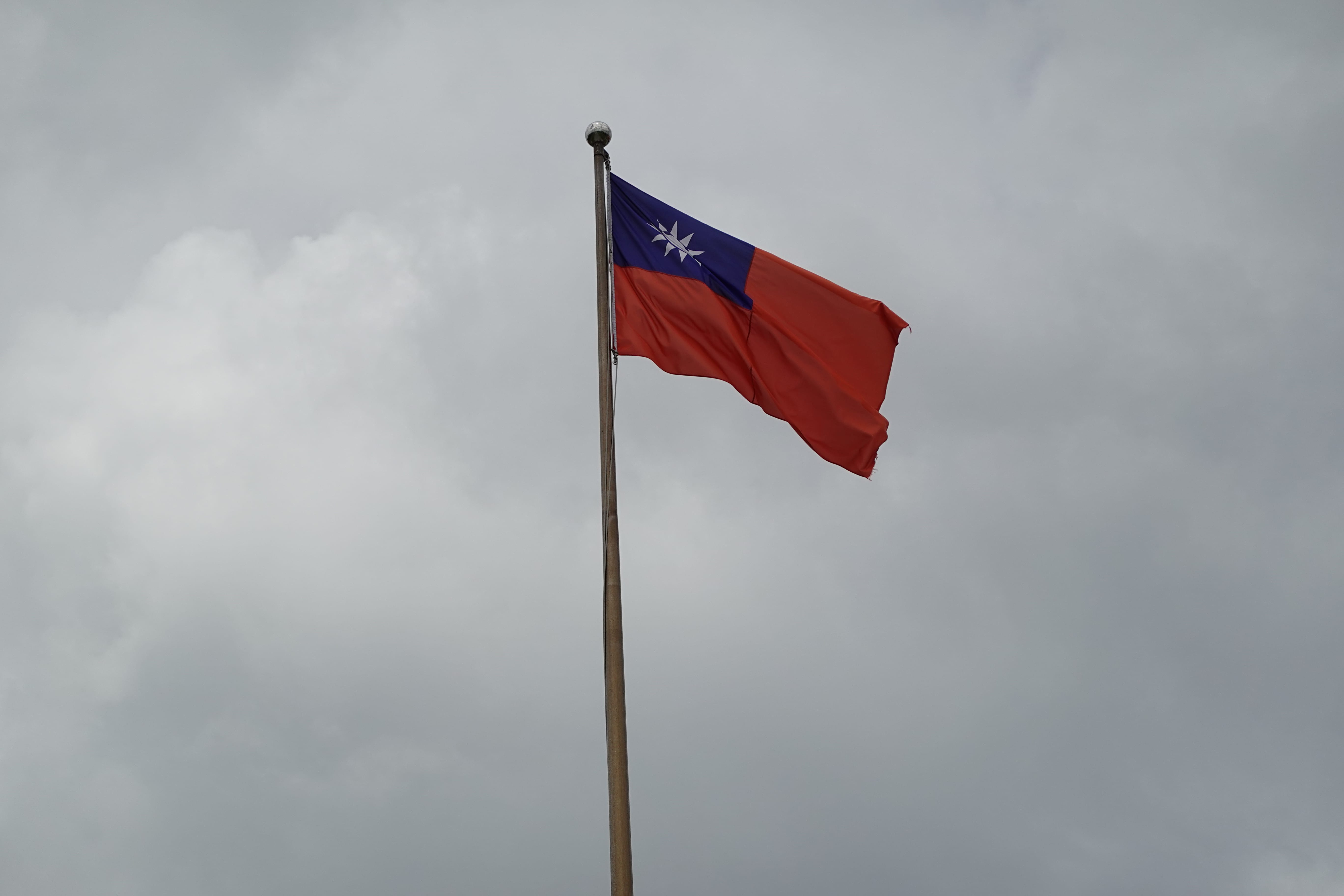 Taiwan flag—blue canton with white sun over a red field—waving on a pole against overcast clouds.