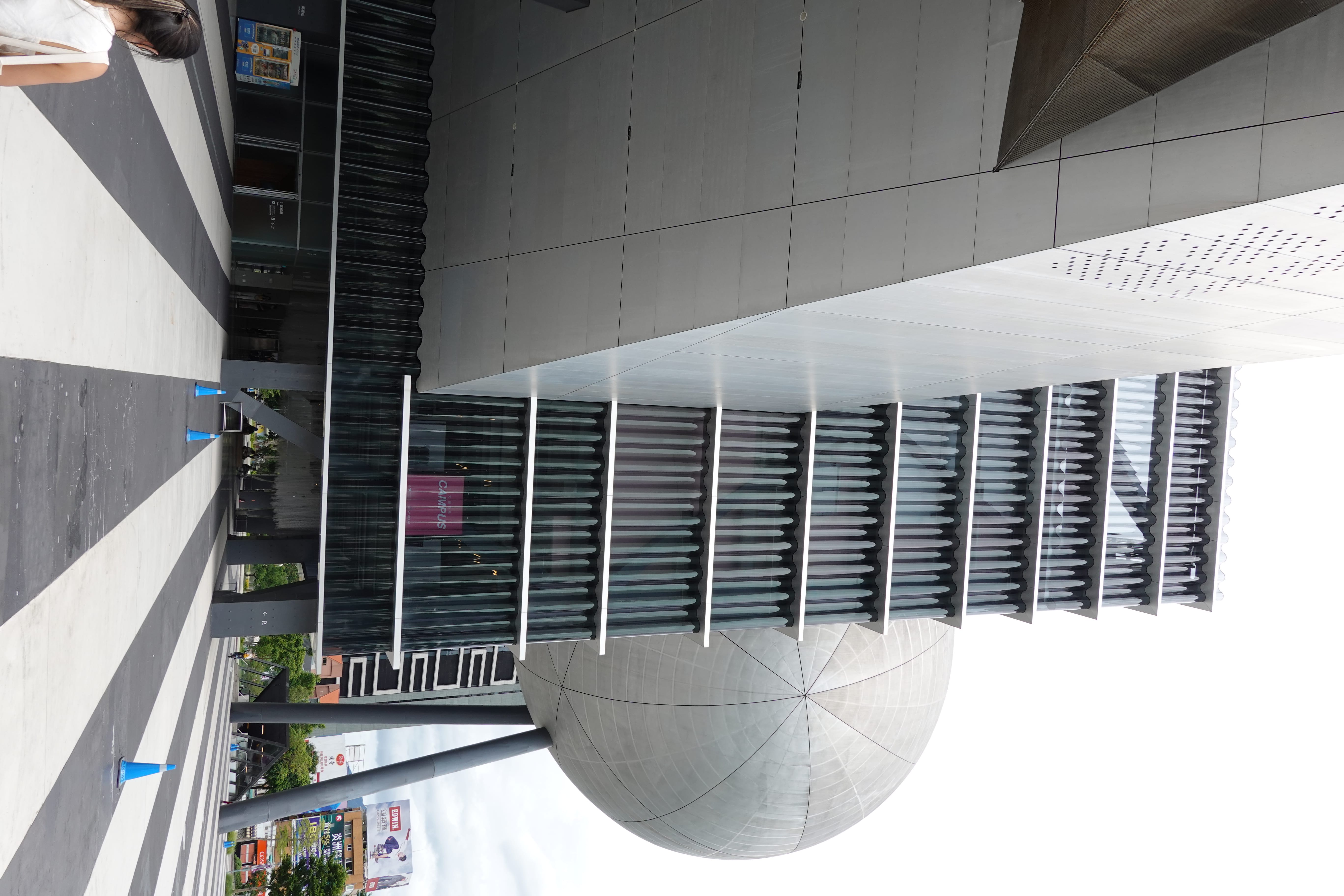 Science museum exterior with a ribbed glass façade and a large silver spherical theater on stilts above a striped plaza. loading=