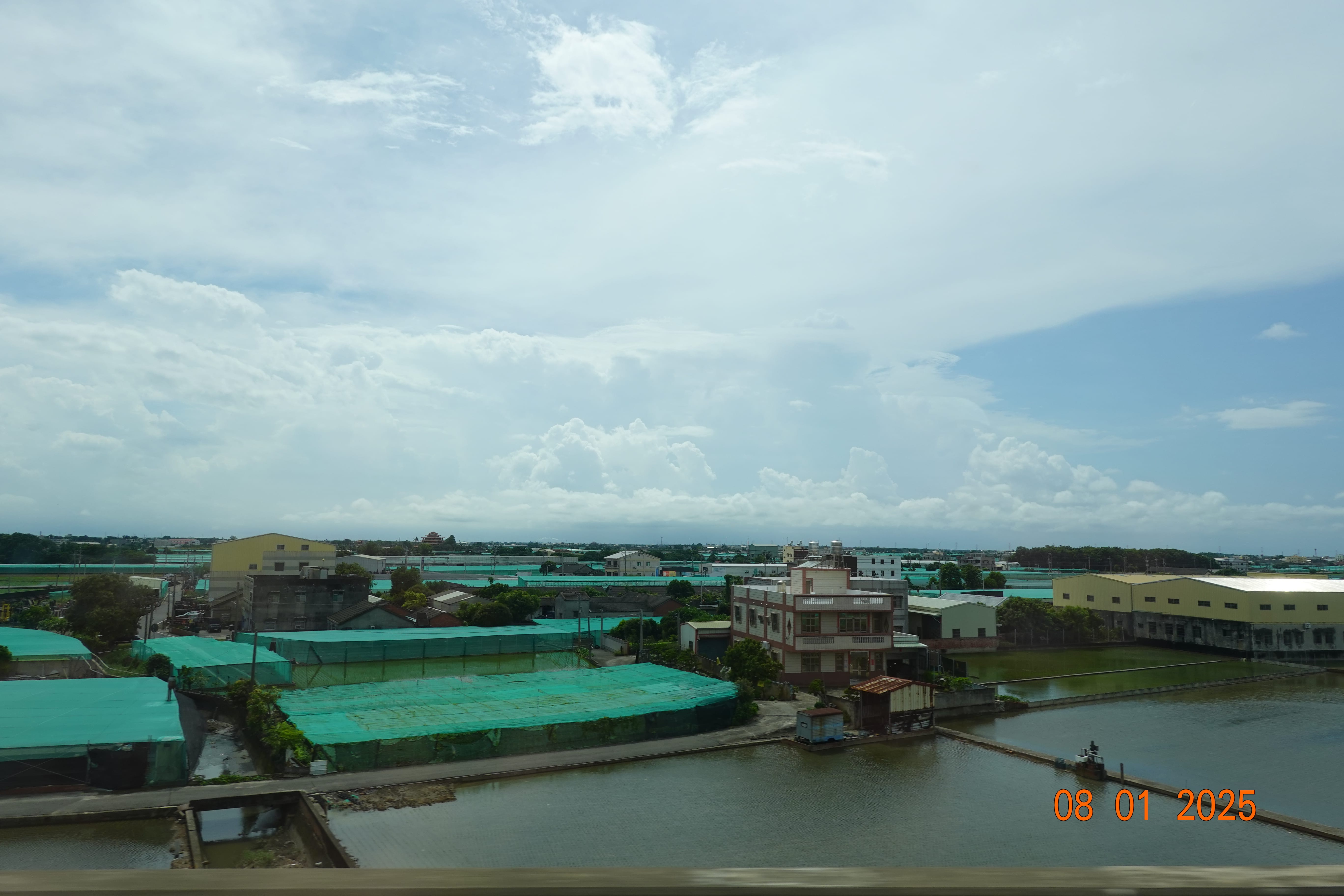 Daytime view from the train: fish ponds and buildings with green tarps under a broad cloudy sky; orange date stamp 08-01-2025.