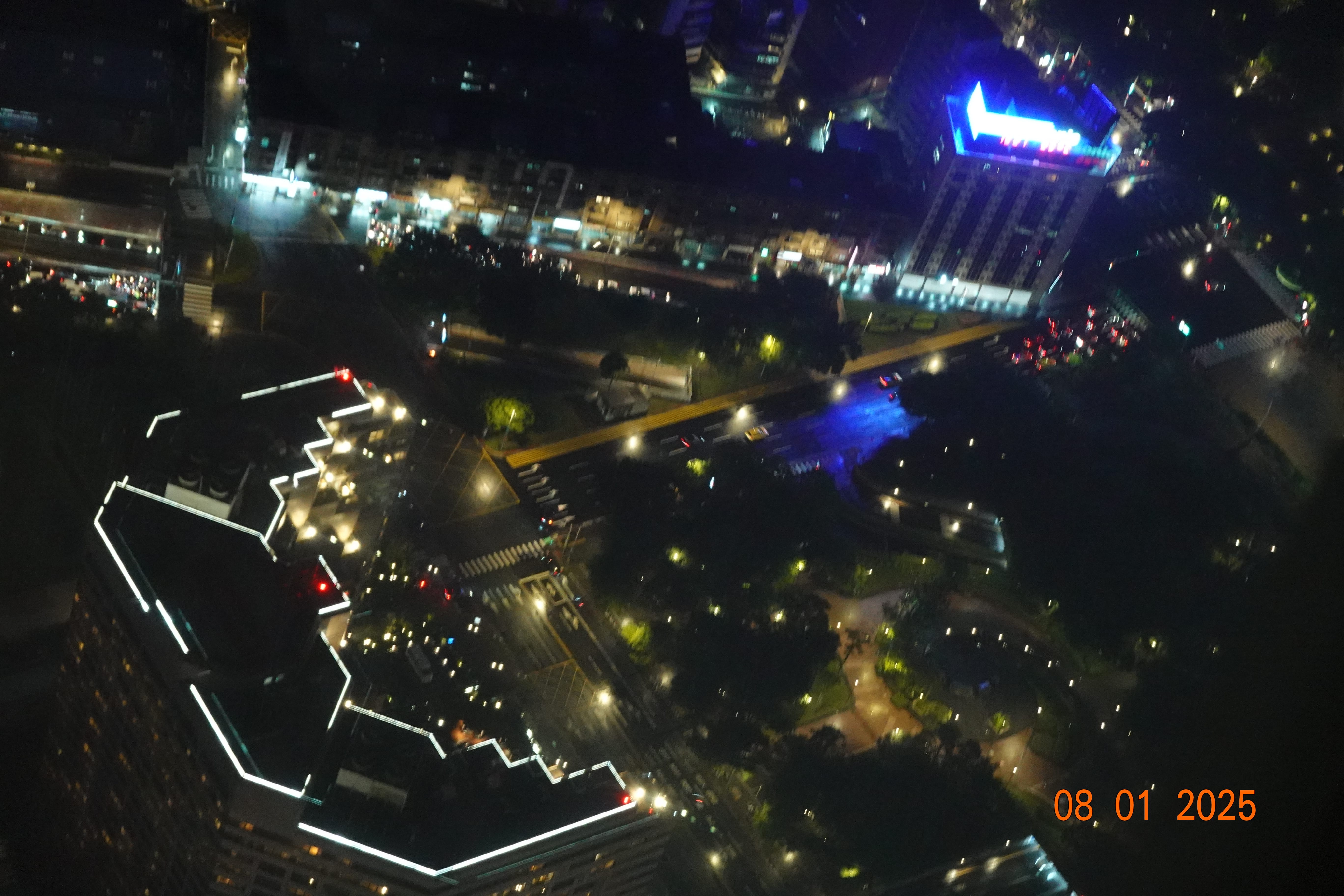 Closer overhead night view of Taipei—streets, park paths and a blue-lit rooftop sign—captured from the observation level; date stamp 08-01-2025.
